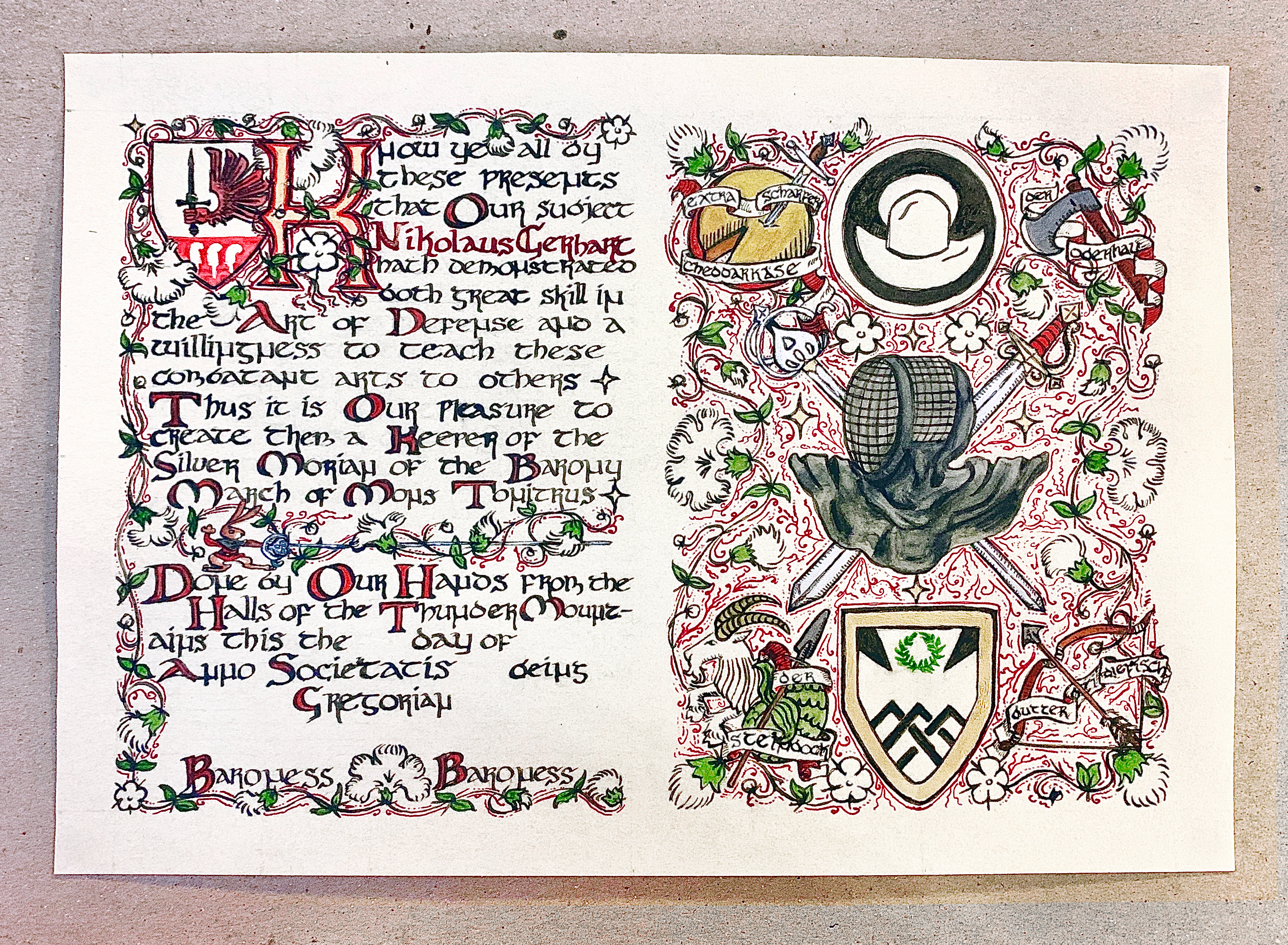 A medieval scroll award in progress: the color stage, red has been added to the left shield device, with gold and red in the capital letter K and some text, red ink decorated vines connect white flowers with green leaves on the left and right side, gray added to the rapier's helmet and many of the weapons, german words have been inked onto scroll-like imagery curling around the artwork on the right. The final stage includes whitework (highlights) on the artwork.