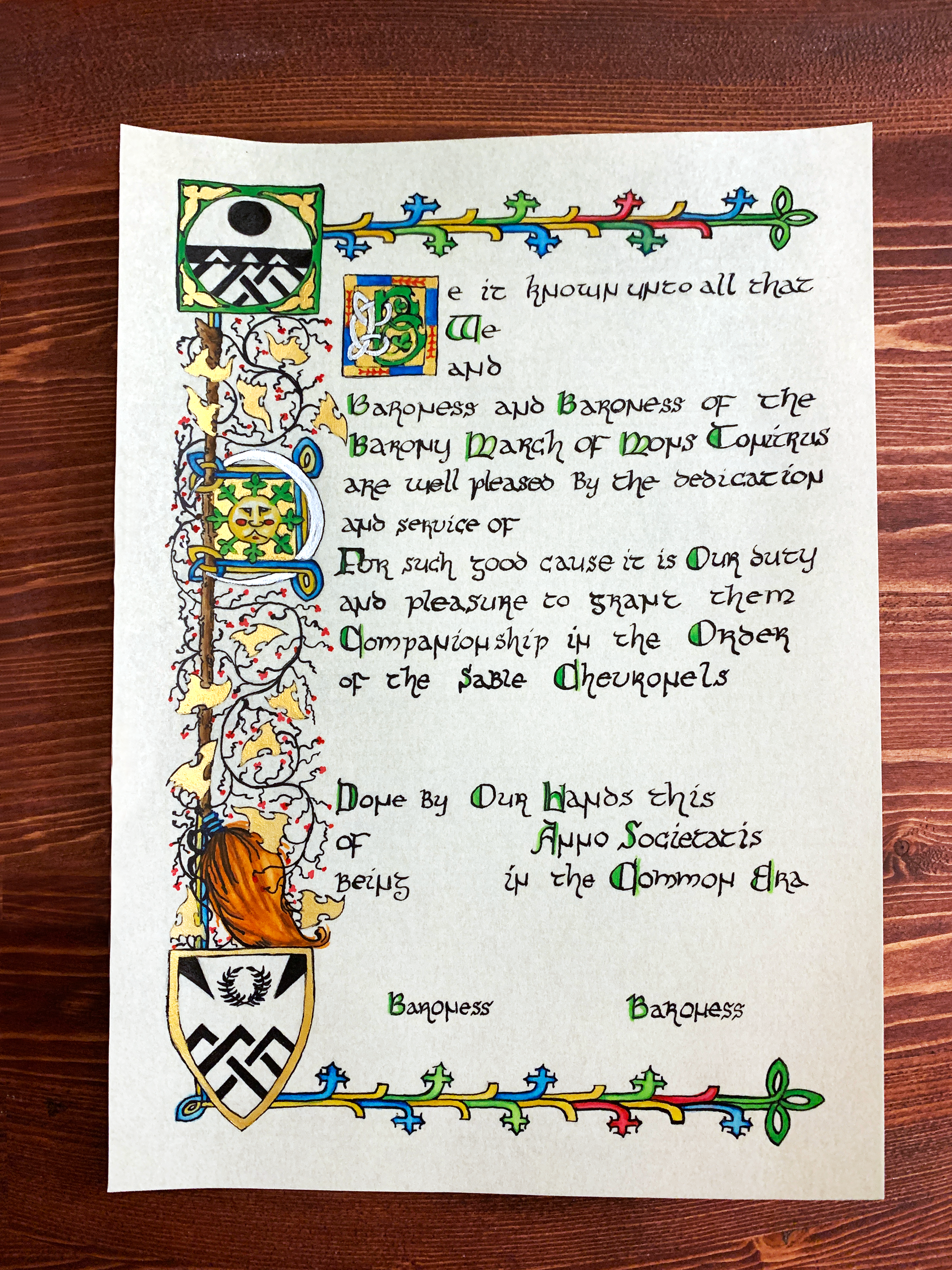 A medieval scroll award in progress: the coloring stage, watercolor has been added to the artwork in the margins, with some capital letters colored green, gold acrylic added to the leaves and borders of shield devices, and silver added to the crescent moon and decorated capital B